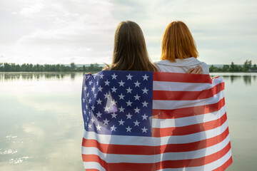 Two young friends women with USA national flag on their shoulders standing together outdoors on lake shore. Patriotic girls celebrating United States independence day.