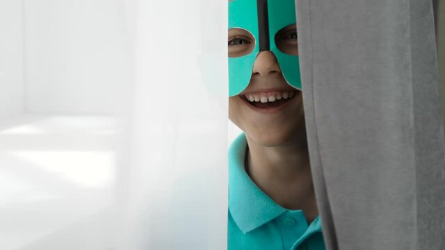 A small charming boy plays hide-and-seek in the room, imagining the hero, wearing a comic mask on his face. Happy child.