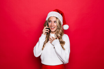 Happy cute woman in santa hat talking on cell phone over red background