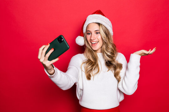 Portrait of a pretty woman wearing Christmas hat taking a selfie isolated over red background