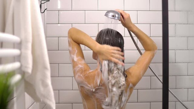 Back view of naked woman washing hair with shampoo while taking a shower