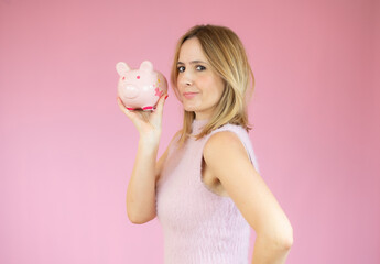 Woman looking at camera holding a piggy bank on her hand against a pink background