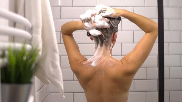 Back view of naked woman washing hair with shampoo while taking a shower