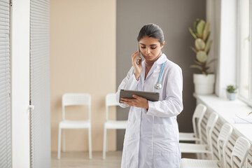 Young female doctor in white coat talking on mobile phone and using digital tablet for online work standing at the corrior at hospital - obrazy, fototapety, plakaty