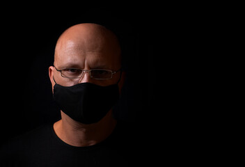 Serious bald middle-aged man with glasses, with a black medical mask from covid 19, on his face, on a black background, with a place for text