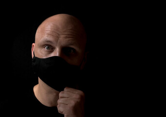 Portrait of surprised middle aged bald man taking off covid 19 black medical mask on black background with space for text