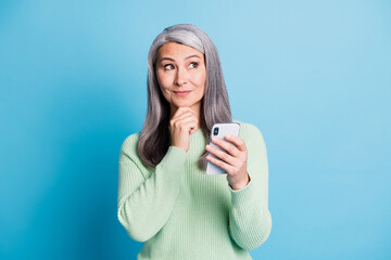 Photo of curious senior woman use phone touch chin dressed green pullover look empty space isolated on blue color background