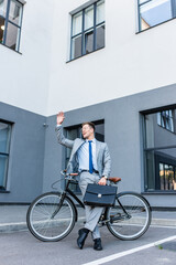 Fototapeta na wymiar Cheerful businessman waving hand while holding briefcase near bicycle outdoors