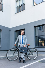 Fototapeta na wymiar Businessman in eyeglasses and suit holding briefcase near bike and building