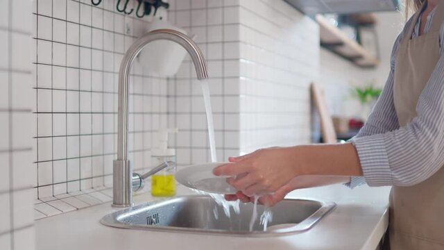 Woman housewife in apron washing plate under running water in sink at modern kitchen