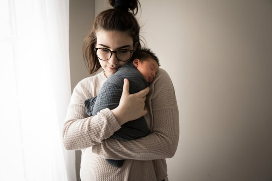 Hipster Millennial Mom Snuggles Swaddled Newborn Son Next to Window