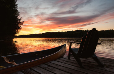 Canoe and adirondack chair on a dock on a lake at sunrise.