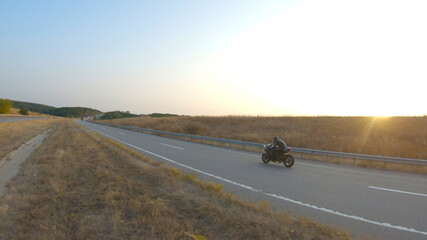 Fototapeta premium Aerial shot of biker is riding on modern sport motorbike at autumn highway. Motorcyclist racing his motorcycle on country road at sunset. Man drive bike during trip. Concept of freedom and adventure