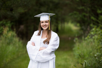 Smiling Female Graduate Wearing White Cap and Gown on Wooded Path