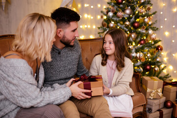 Christmas and family concept - parents giving present to their daughter in living room with decorated Christmas tree