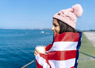 Beautiful young woman standing wrapped in the American flag with a cup