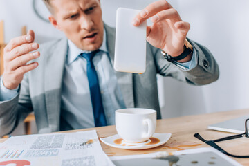 Confused businessman holding dirty smartphone near pouring out coffee and papers on table