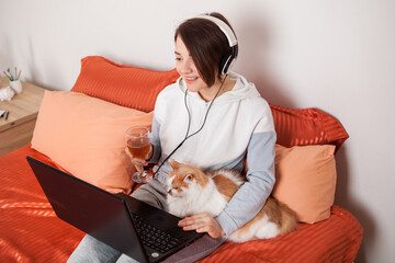 a girl with a glass of wine communicates online with friends and relatives. A woman with a laptop on the bed and a cat communicates by video link