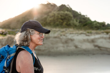 Active older woman wearing a backpack and hat walking on a beach in New Zealand