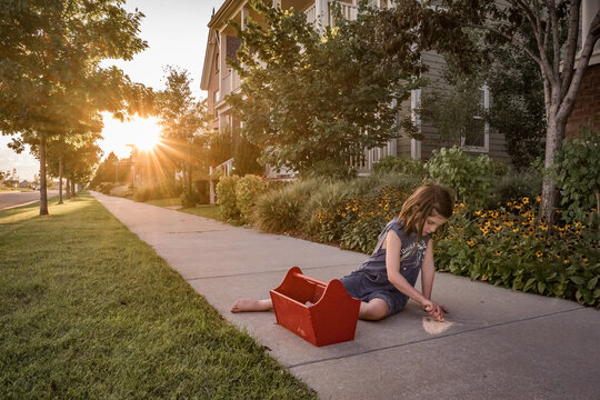 young girl draws with sidewalk chalk in front of house at sunset
