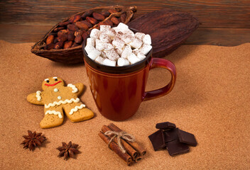 Cup of cacao drink with marshmallow, gingerbread man, chocolate, spices, cocoa pods and cocoa beans on a cork background.