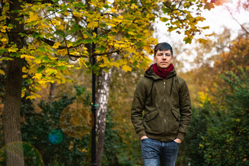 Young man in a red scarf, green jacket and blue jeans looking at the camera with his hands in his pockets against a background of yellow foliage. Object position to the right