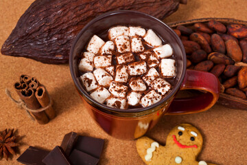 Cup of cacao drink with marshmallows, gingerbread man, chocolate, spices, cocoa pod and cacao beans on a cork background. Winter drink concept.