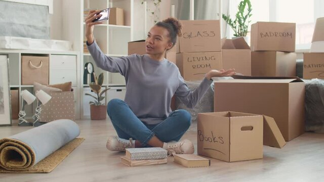 Lockdown of young curly-haired woman sitting on floor with smartphone in hands and shooting video while packing things into cardboard boxes for move