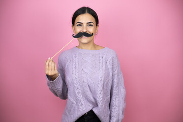 Beautiful woman wearing a casual violet sweater over pink background having fun wearing a fake mustache.