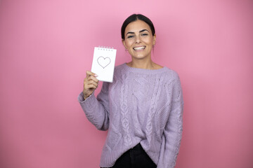 Fototapeta na wymiar Beautiful woman wearing a casual violet sweater over pink background holding notebook with a hart message