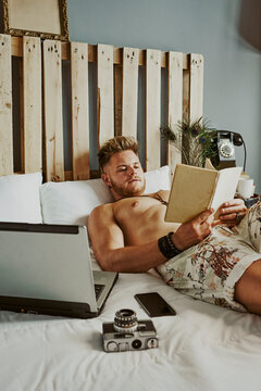 A man reads a book while working with his cell phone and laptop in a hotel bed