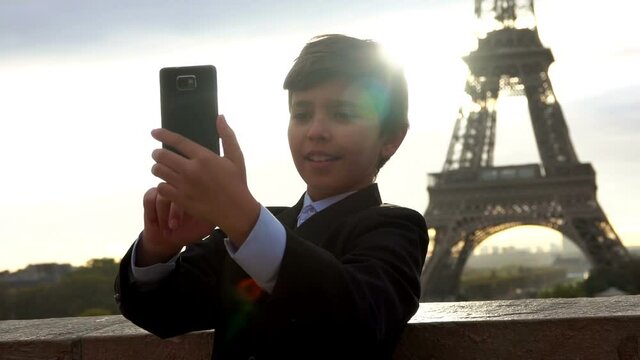 A happy teenage boy in a black suit is taking a photo on the phone on the background of the Eiffel tower at the sunrise, Paris, France