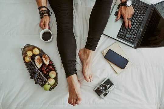 Feet and legs of a man in bed next to a computer, fruit, coffee,