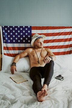 A young man in bed with a hat and no shirt with the American fla