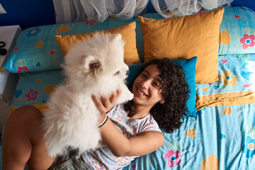 A little girl is playing with her white Pomeranian puppy in bed.
