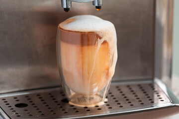 Coffee streaming from coffee machine and pouring out of glass full of milk foam.