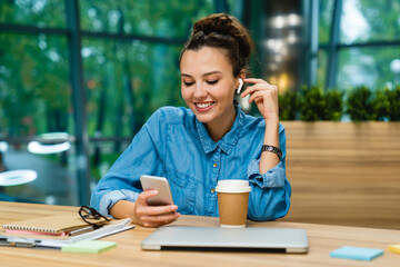 Charming young busy girl checking her phone with earbuds sitting at the desk with laptop and cup of coffee