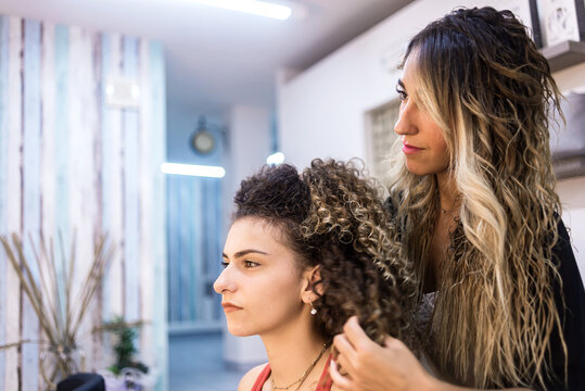 Hairdresser combing client with long curly hair against wall mirror at Hair Salon