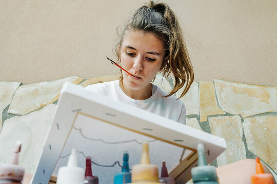 white girl biting a bush while painting with her finger her picture in front of bottle paintings. Overhead horizontal photo