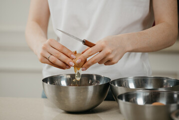 A close photo of the hands of a girl who is breaking the organic farm egg holding a knife above the stainless steel soup bowl in the kitchen.