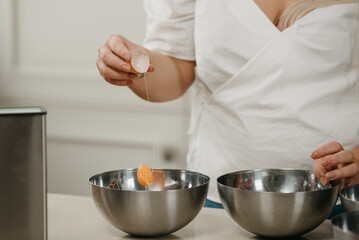 A photo of the hand of a young woman who is dropping the yolk of the organic farm egg to the stainless steel soup bowl in a kitchen.