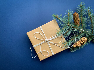 Homemade gift box wrapped in craft paper next to fir tree branches. Top view holiday, winter blue background with copy space.