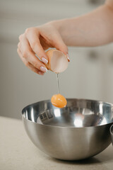 A close photo of the hand of a woman who is dropping the yolk of the organic farm egg to the stainless steel soup bowl in a kitchen. A falling yolk.