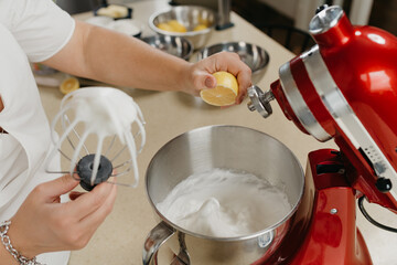 A photo of the hand of a young woman who is crushing lemon juice to the mixed meringue in a stainless steel soup bowl in a kitchen.