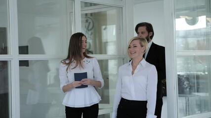 Team of young promising businessmen come into the office and laugh. A man in a black suit and two women go and discuss work issues. - Powered by Adobe