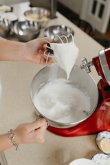 A close photo of the hand of a young woman who is demonstrating meringue on the mixer whisk near a stainless steel soup bowl in a kitchen.