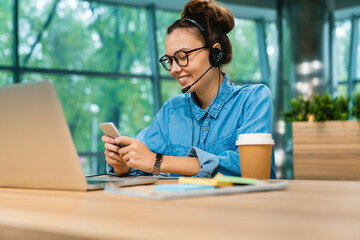 Smiling confident businesswoman checking her phone with headset and laptop at office desk