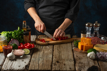Professional chef cuts with knife cherry tomato on wooden chopped board for cooking tasty food for dinner. Backstage of preparing meal on rustic table with ingredients on dark blue background.