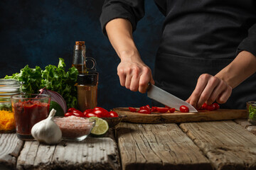 Professional chef cuts with knife cherry tomato on wooden chopped board for cooking tasty food for dinner. Backstage of preparing meal on rustic table with ingredients on dark blue background.Close-up