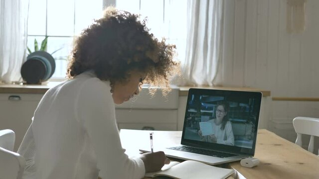 Concentrated African-American girl attends online lesson sitting against laptop and writing abstract close view slow motion